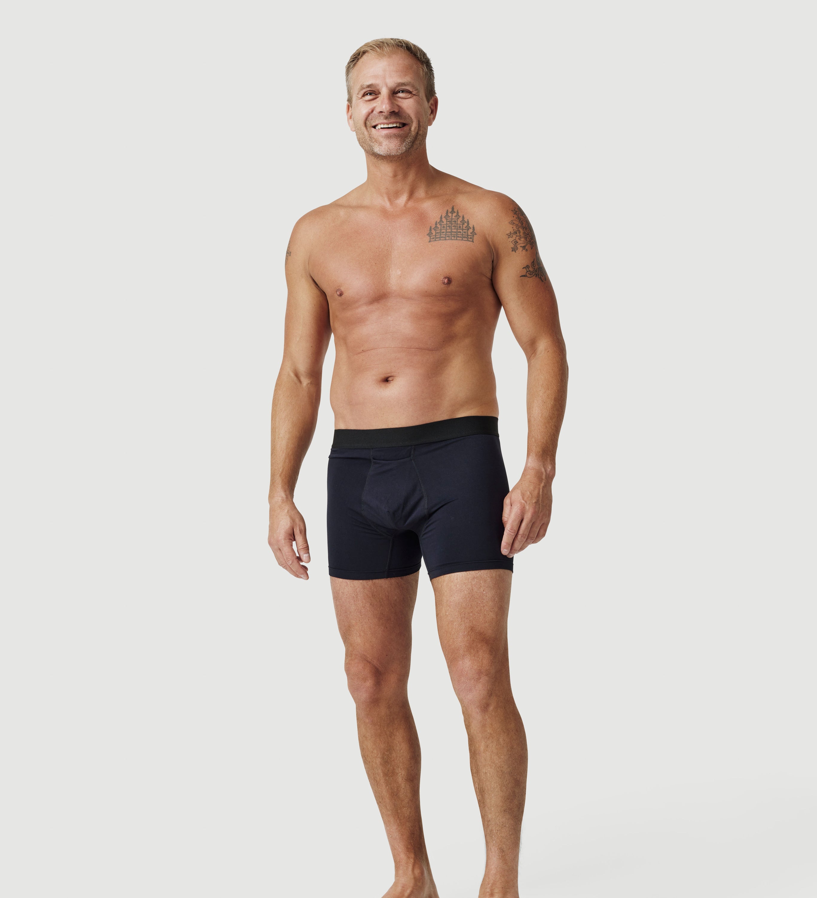 Smiling man in black boxer briefs with tattoos on upper body, standing on white background